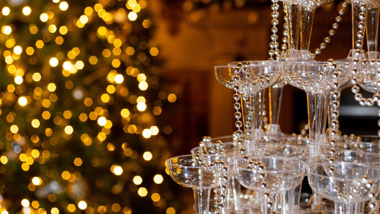 A pyramid of champagne glasses, decorated with strings of pearls. In the background the lights of a Christmas tree.